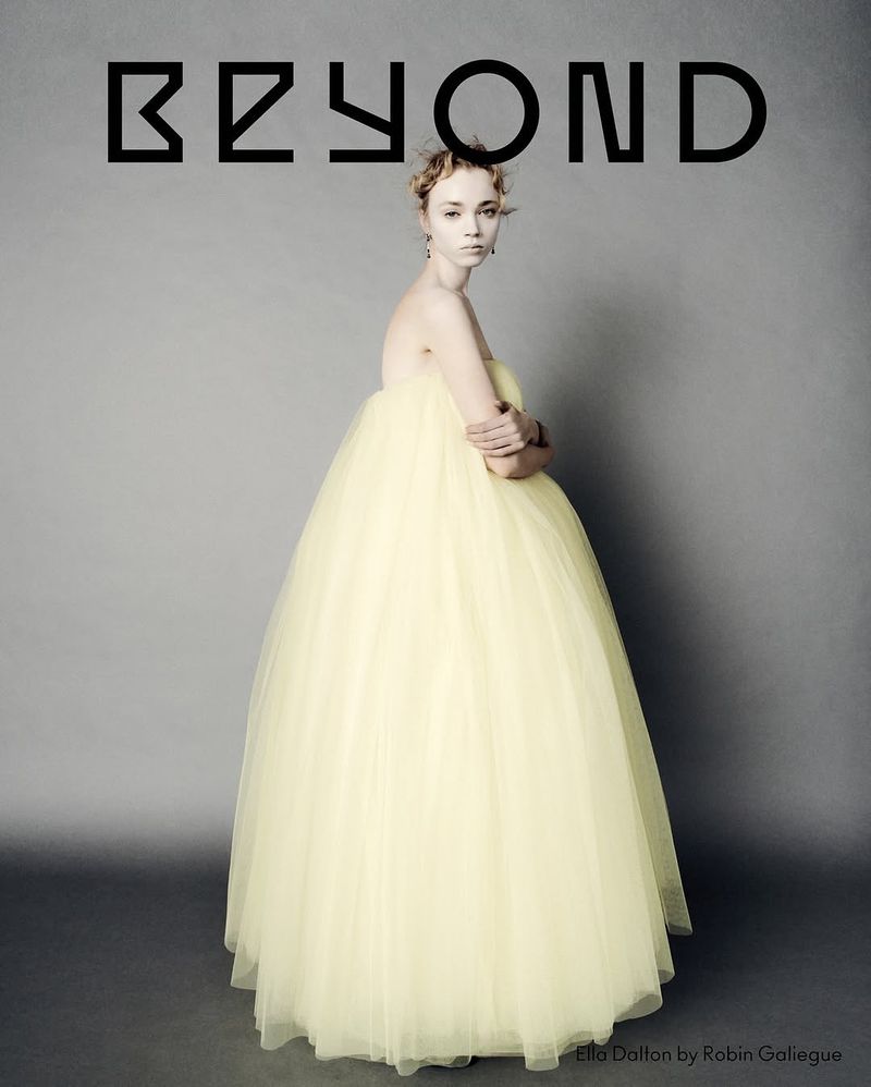 Portrait of a slender woman in a formal pale yellow tulle gown with arms crossed against a grey backdrop with the word BEYOND above her.
