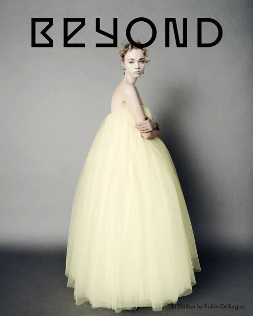 Portrait of a slender woman in a formal pale yellow tulle gown with arms crossed against a grey backdrop with the word BEYOND above her.