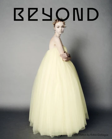 Portrait of a slender woman in a formal pale yellow tulle gown with arms crossed against a grey backdrop with the word BEYOND above her.