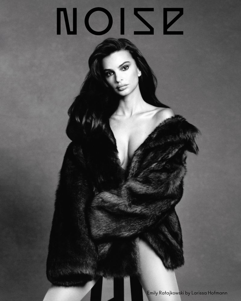 Emily Ratajkowski poses in a fur coat for a black and white portrait. The model is sitting on a stool in front of a gray background.