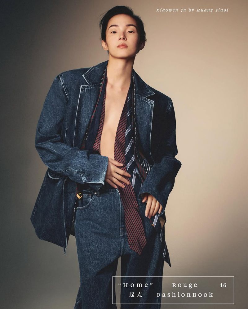Portrait of a slender Asian woman wearing a denim blazer, jeans, and a striped tie, posing in a studio with soft lighting.