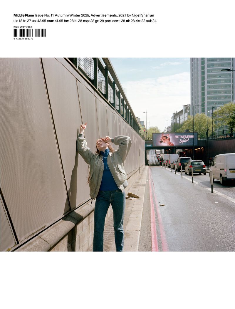 Adult female stands near a wall on a city street, wearing a jacket and jeans, with city buildings and a billboard in the background.