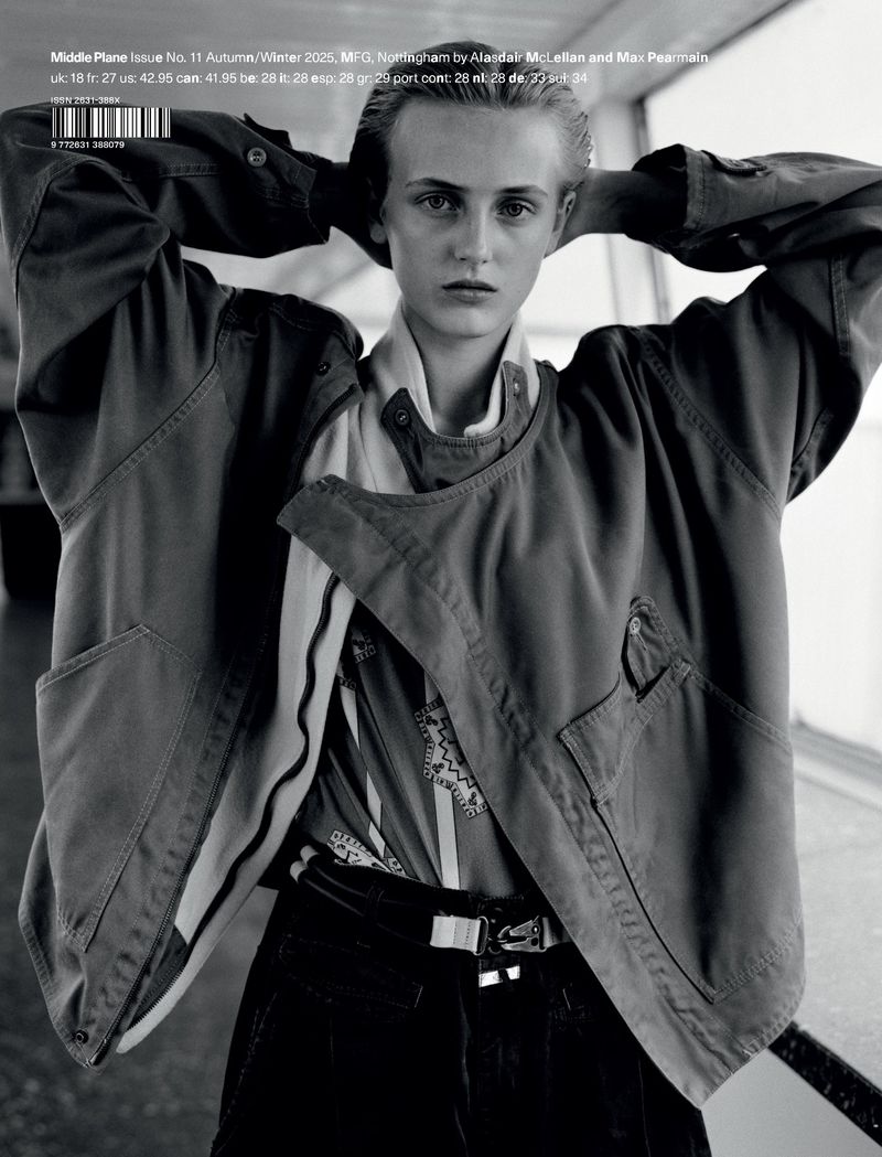 Monochrome fashion shot of a young fair-skinned person in a jacket, graphic shirt, and black pants posing against an indistinct interior backdrop.