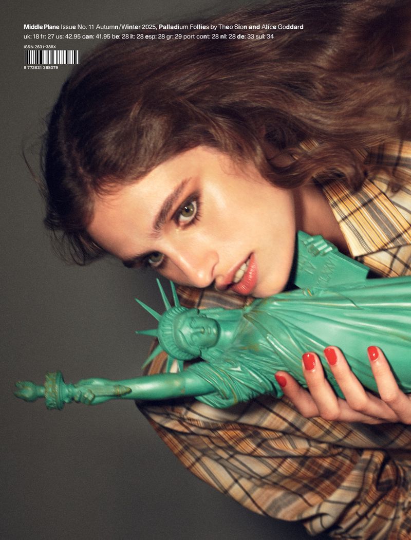 Brunette woman in plaid shirt holding Statue of Liberty figurine in studio with soft lighting for fashion or celebrity editorial.