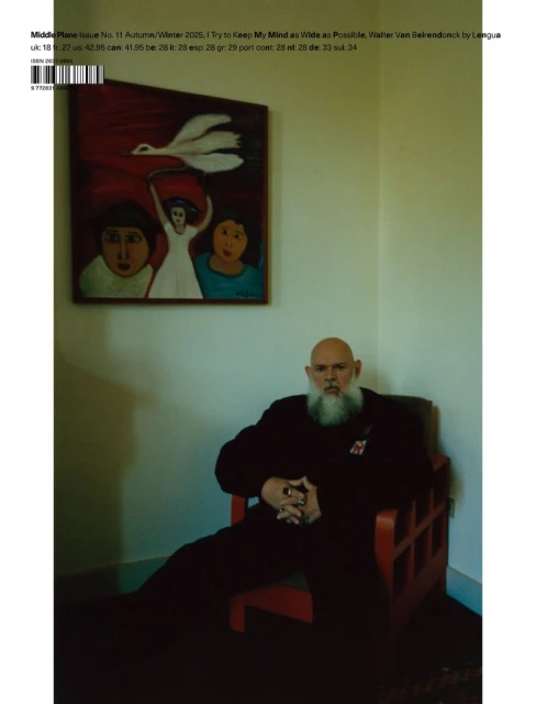 Elderly man with a long white beard sits in a red chair with a painting on the wall, creating a contemplative portrait.