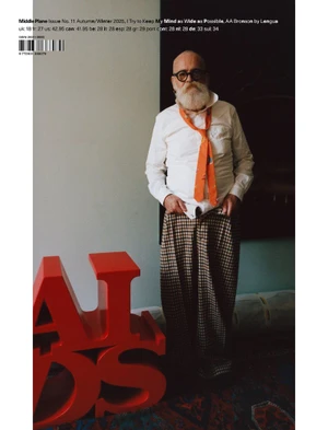 Artist AA Bronson wearing an orange tie and houndstooth trousers standing beside a red AIDS sculpture for Middle Plane Magazine.
