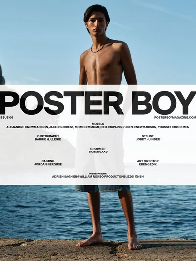 Shirtless male model wearing denim shorts standing barefoot on a stone ledge by the ocean on a magazine cover.