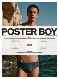 A young male model standing shirtless against a rocky coastal background with dark water and clear blue sky.