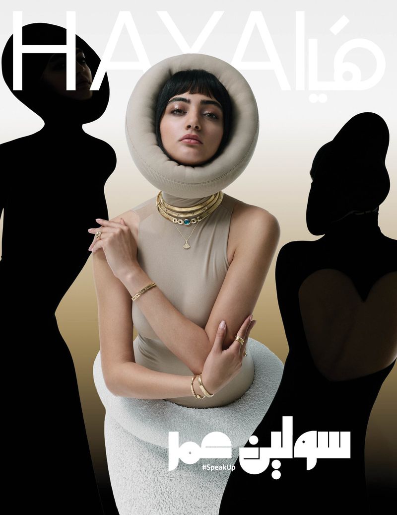 Magazine cover featuring a woman with dark hair and statement jewelry, seated in a uniquely designed chair, flanked by silhouette figures.