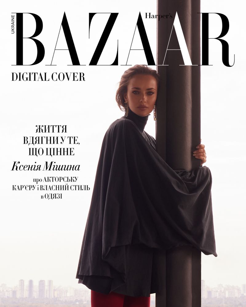 Fashion editorial cover featuring an adult woman in a black poncho posing by a dark pillar in front of a cityscape, with Bazaar logo.