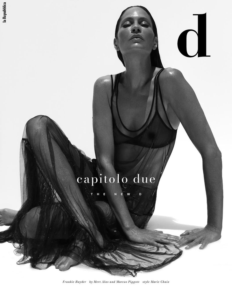 Black and white portrait of an adult woman in a sheer black dress sitting against a white backdrop in a fashion editorial style.