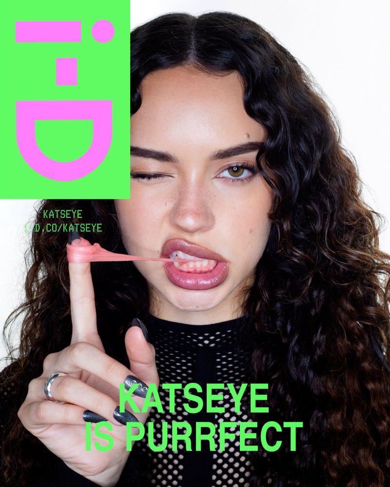Portrait of a young woman with brown curly hair winking with bubblegum on her tongue, wearing a black mesh top with pink and green design.