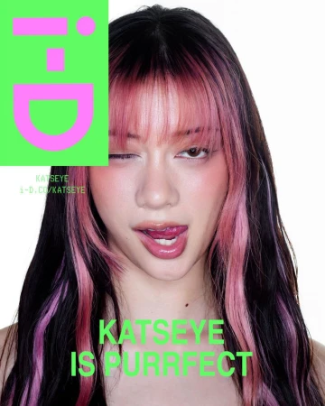 Close-up portrait of a woman with pink-streaked dark hair winking and sticking out her tongue against a white seamless background.
