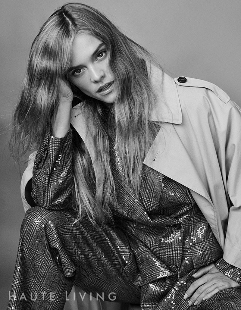 Monochrome portrait of a woman with long hair wearing a trench coat and patterned pants looking at the camera with a hand to her head.