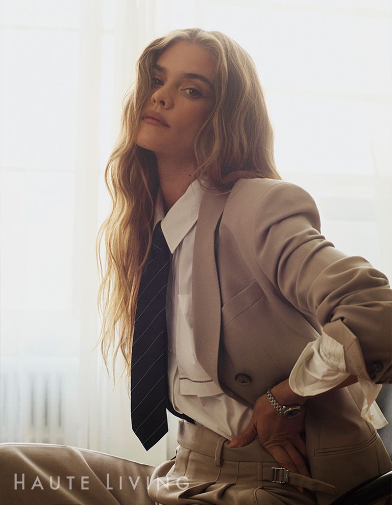 Portrait of a fair-skinned adult woman with long blonde hair wearing a beige blazer, white shirt, and dark tie indoors with soft window lighting.