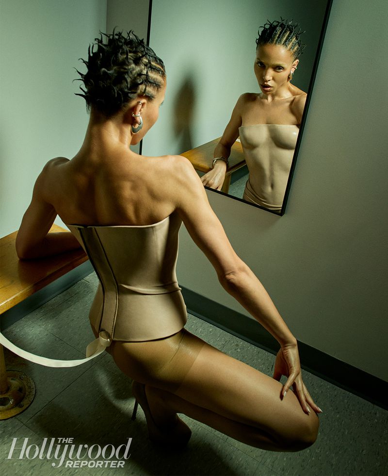 Adult woman in a beige corset and tights is seen kneeling next to a bench as she looks into a mirror in an indoor setting.