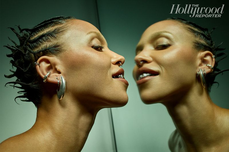 Close-up profile of a woman with braided hair, nose ring, ear jewelry, and grill, reflected in a mirror with logo in the background.
