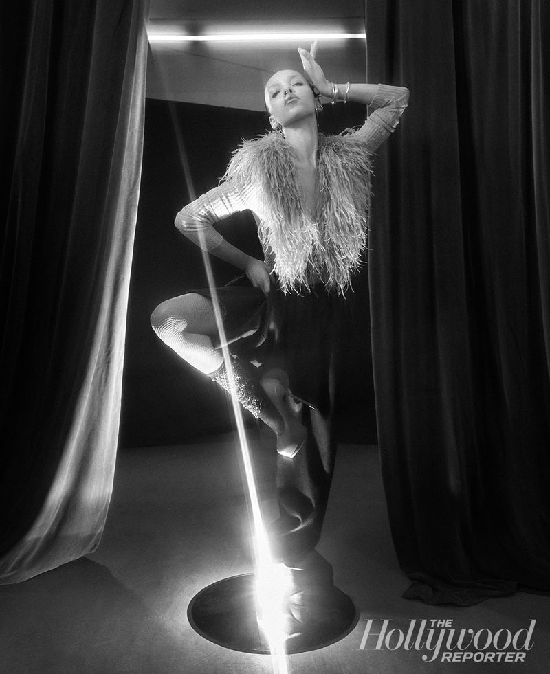 Black and white portrait of a bald woman wearing a feather-trimmed top, posing confidently in front of a spotlight and dark curtains.