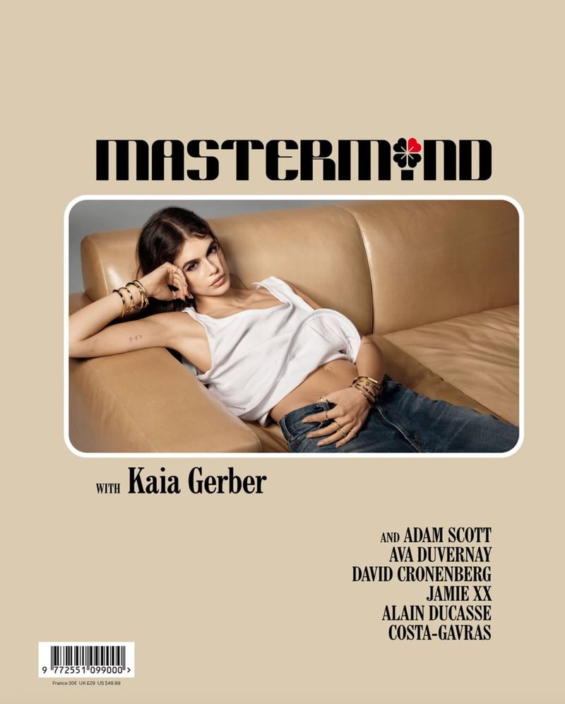 Indoor portrait of model Kaia Gerber reclining on a tan leather sofa, wearing a white tank top and blue jeans against a tan background.