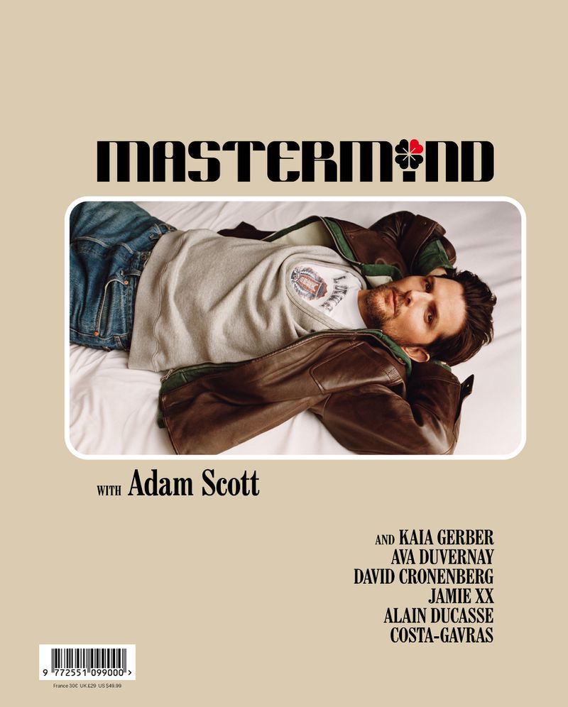 Adult male, Adam Scott, lies on white bedding wearing blue jeans, a cream-colored shirt, and a brown leather jacket in a magazine cover shot.