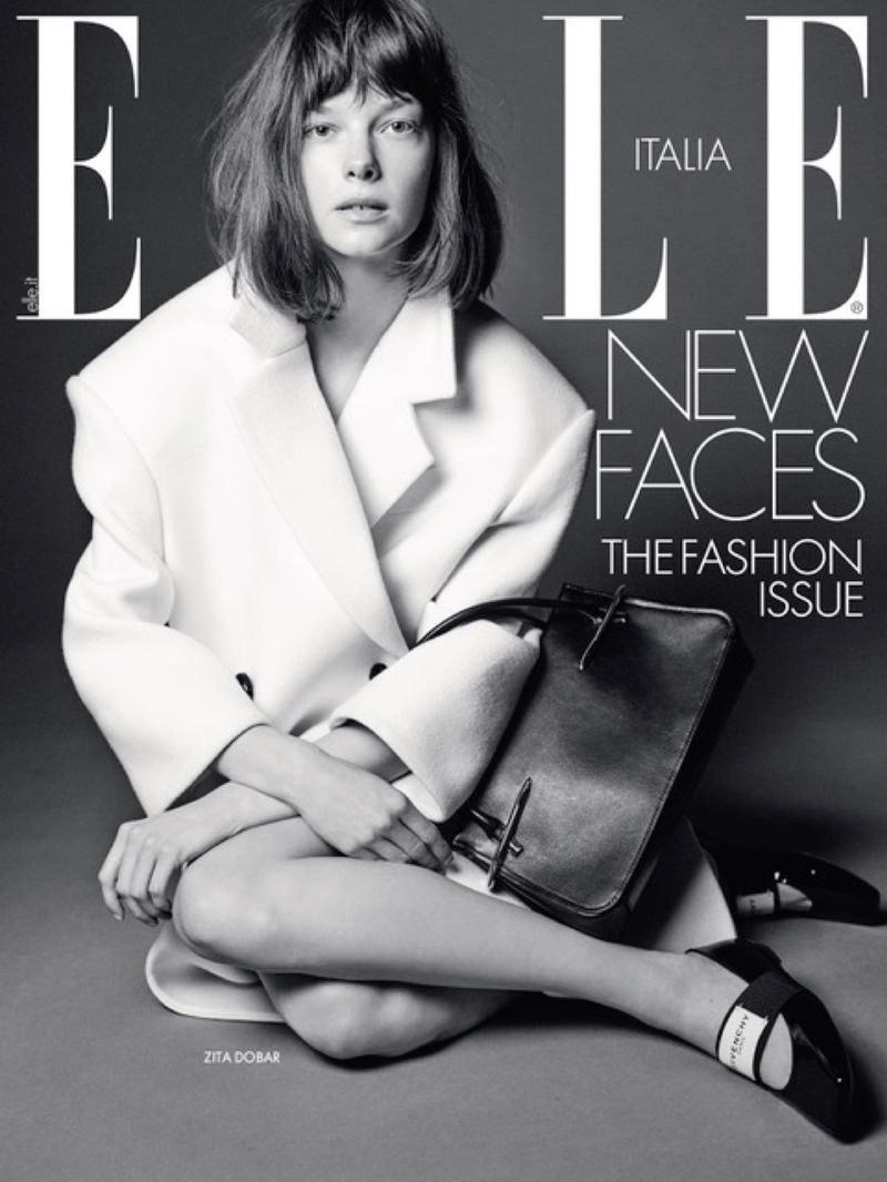 Elle Italia magazine cover featuring a young woman in a white coat, seated with legs crossed, holding a black bag, and wearing black shoes.