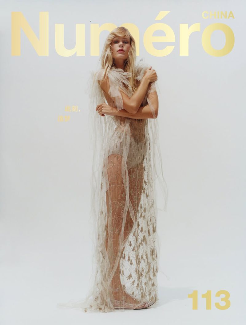 Fashion editorial cover featuring a blonde fashion model wearing sheer lace dress in a full shot, against a clean white backdrop.