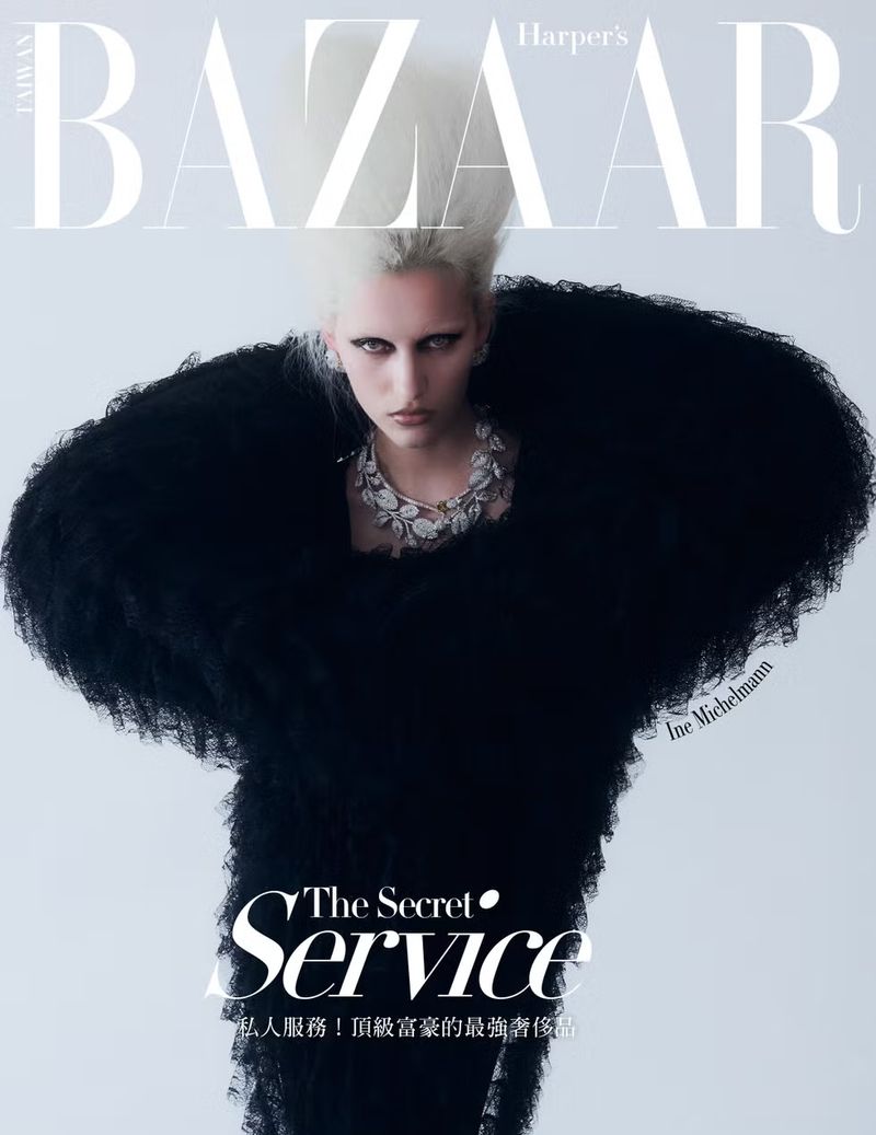 Fashion editorial cover featuring a model with platinum blonde hair wearing an extravagant black dress and statement jewelry in a studio.