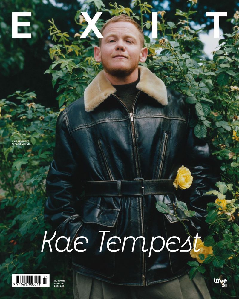 Kae Tempest on the cover of EXIT magazine wearing a black shearling coat, posing outdoors amongst green foliage and yellow flowers.