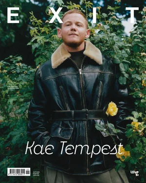 Kae Tempest wearing a belted leather jacket with a shearling collar in front of lush rose bushes with yellow flowers.