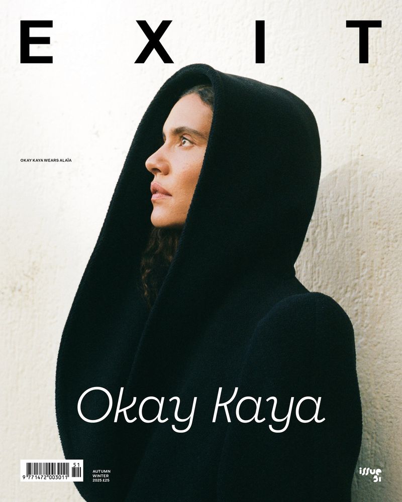 Kaya Wilkins wearing a sculptural black hooded coat in profile against an off-white textured wall for an Exit Magazine cover.