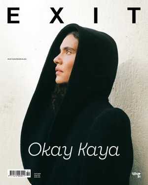 Kaya Wilkins wearing a sculptural black hooded coat in profile against an off-white textured wall for an Exit Magazine cover.