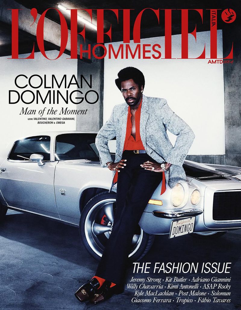 Colman Domingo leans against a classic car for a magazine cover. He wears a suit with an open red shirt and black pants in a studio.