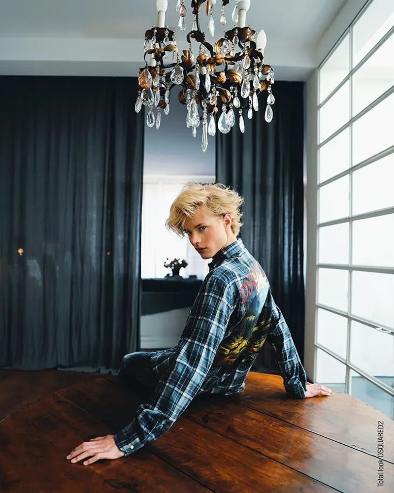 Young blond man poses on a table in a blue plaid shirt beneath a chandelier, indoor shot, fashion editorial, lifestyle photography.