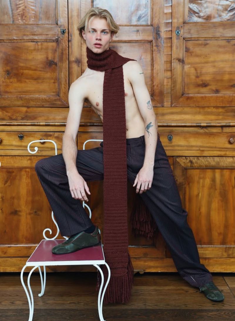 Man with blond hair wearing long brown scarf with pinstripe pants and black shoes sits on white chair in front of brown wooden doors.