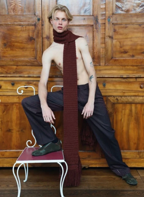 Man with blond hair wearing long brown scarf with pinstripe pants and black shoes sits on white chair in front of brown wooden doors.