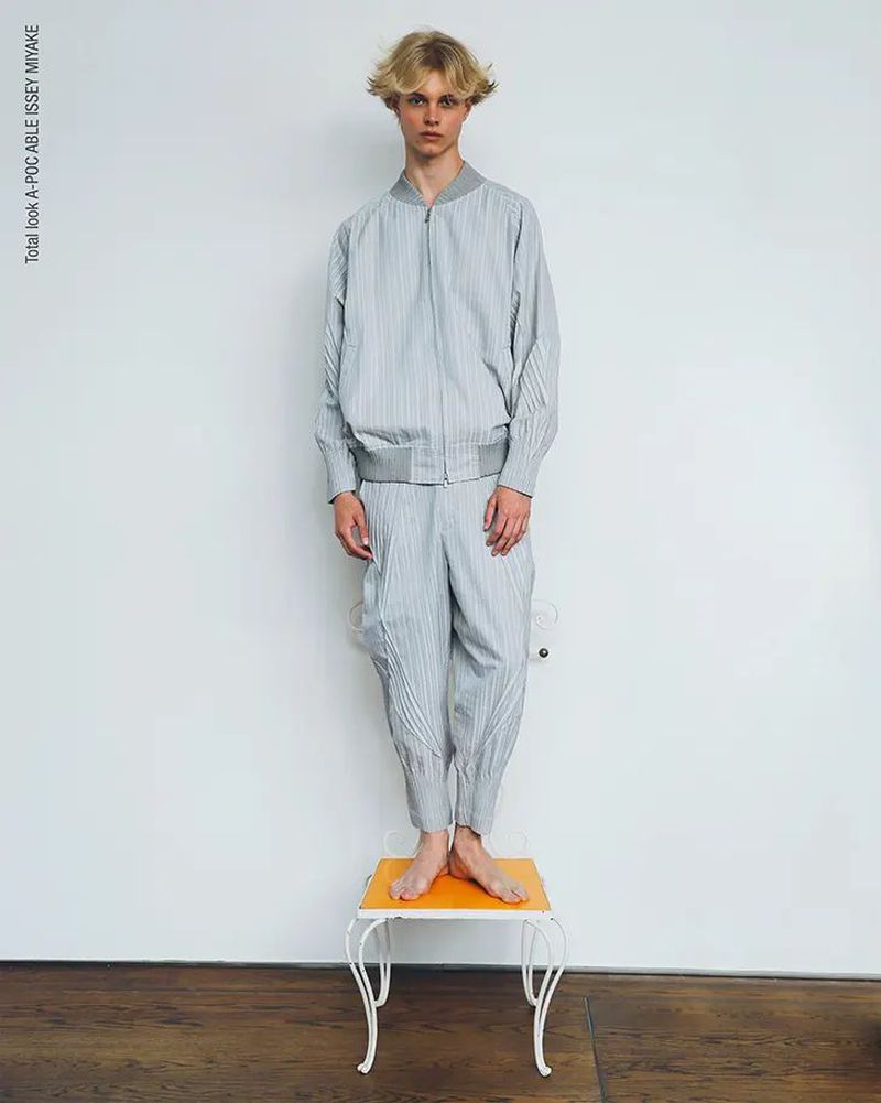 Young adult man in grey striped Issey Miyake outfit stands barefoot on orange table against white wall in a fashion studio shot.