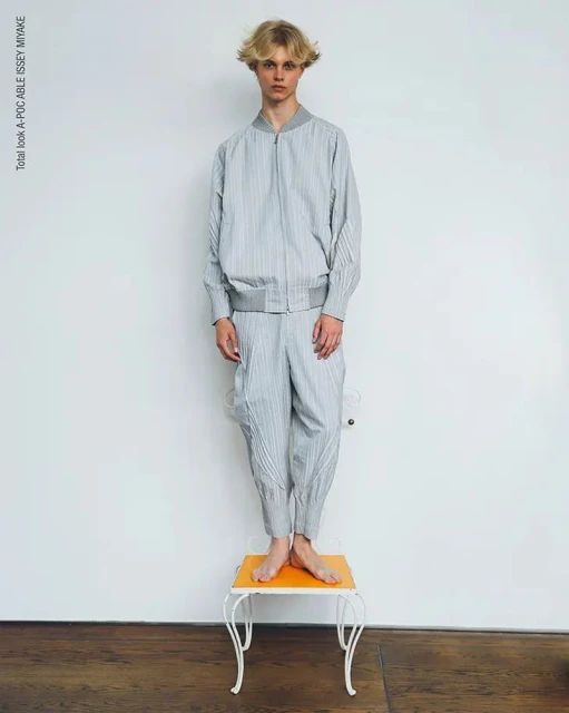Young adult man in grey striped Issey Miyake outfit stands barefoot on orange table against white wall in a fashion studio shot.