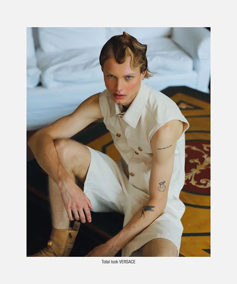 A fair-skinned male model with blonde hair poses indoors. He is wearing a beige, sleeveless shirt and matching shorts, creating a stylish look.