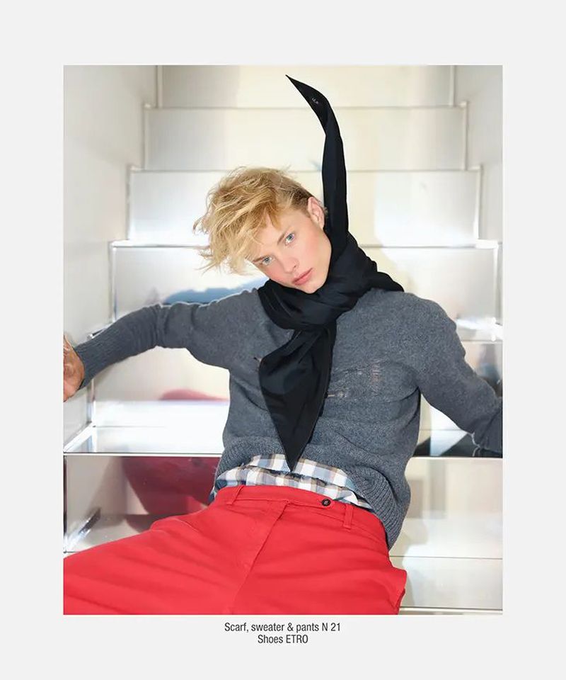 Fashion shot of a blond male model on silver stairs, wearing a gray sweater, bright red pants, and a long black scarf.