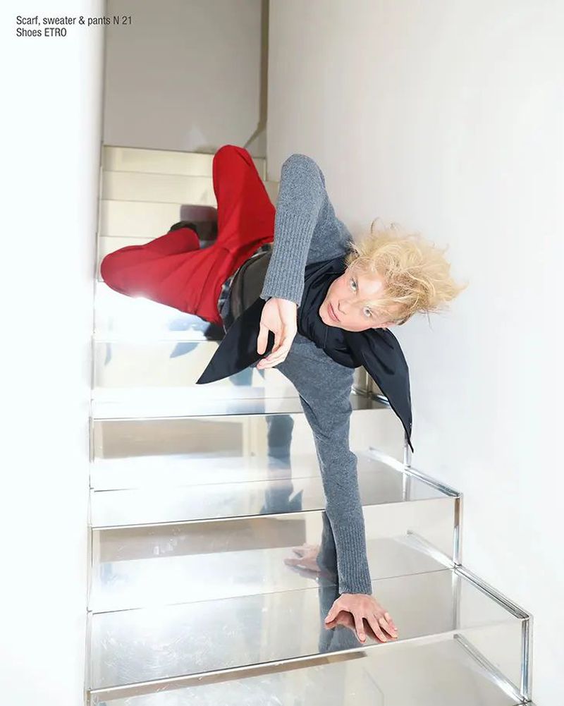 Fashion shot of a young adult with blond hair wearing a grey sweater, black scarf, and red pants posing on silver stairs indoors.