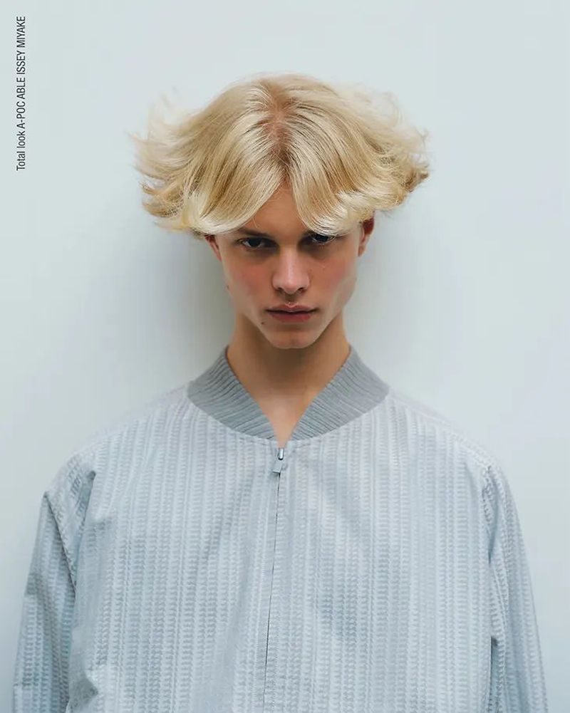 Close-up studio portrait of a young adult with blonde hair, wearing a gray textured bomber jacket, against a white backdrop.