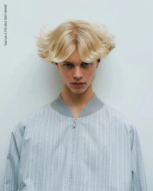 Close-up studio portrait of a young adult with blonde hair, wearing a gray textured bomber jacket, against a white backdrop.