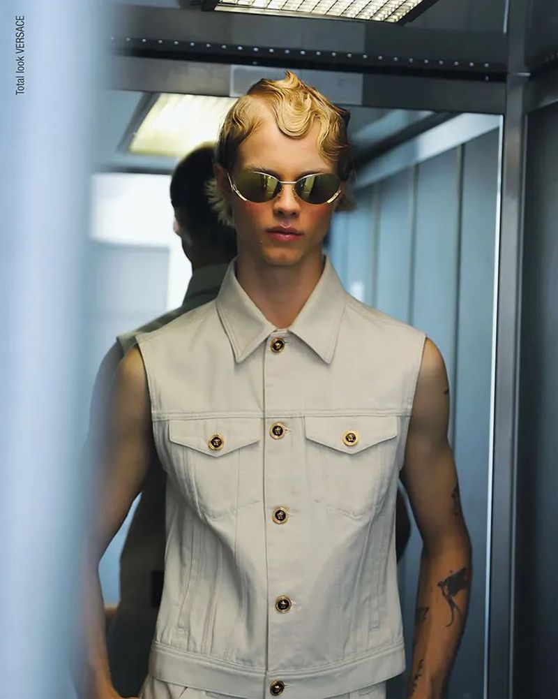 A blonde male model wears sunglasses and a sleeveless beige denim jacket in an indoor setting, posing in front of a mirrored wall.