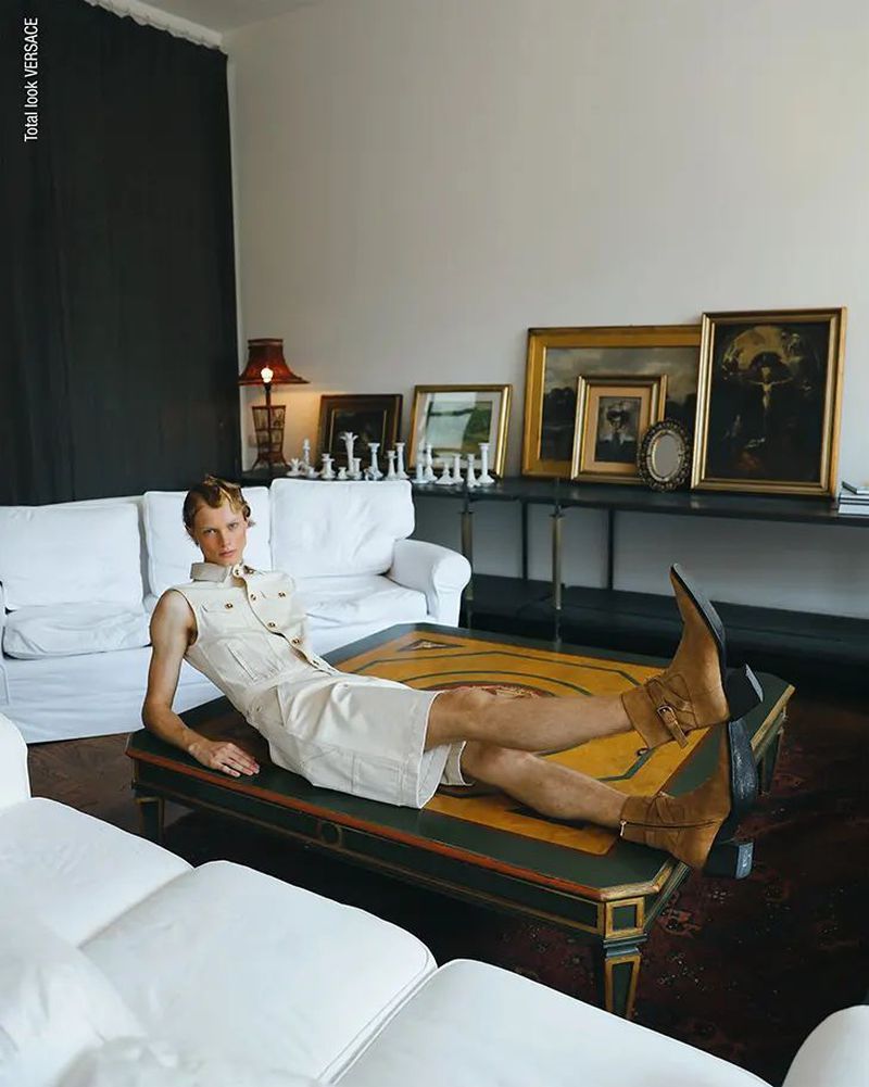A blond male model reclines on a coffee table with western boots, wearing a short, light-beige sleeveless shirt dress in a sunlit room.