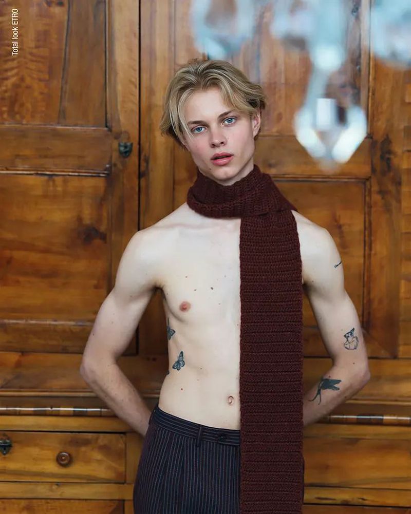 Young adult male poses in front of a wood paneled cabinet wearing a brown scarf and striped pants in a portrait shot.