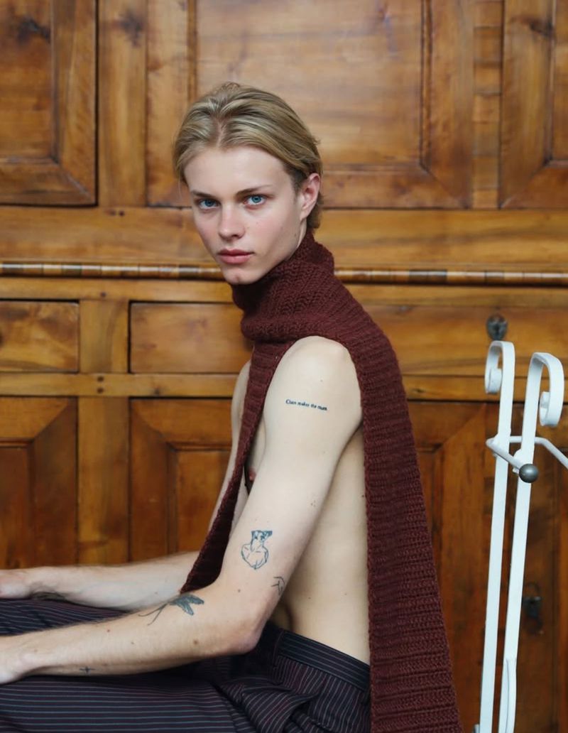 Portrait of a young man with blond hair wearing a brown scarf and striped pants, seated in front of a wooden door.