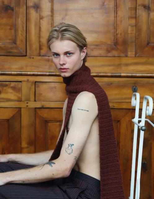 Portrait of a young man with blond hair wearing a brown scarf and striped pants, seated in front of a wooden door.