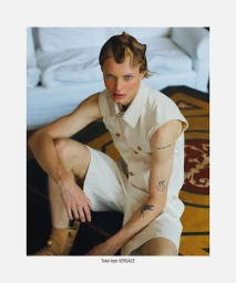 Young man with styled hair and tattoos wearing a light cream sleeveless shirt and shorts, seated indoors.