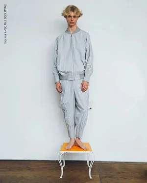 Young male model standing barefoot on a small yellow stool wearing a light grey striped matching bomber jacket and trousers set.