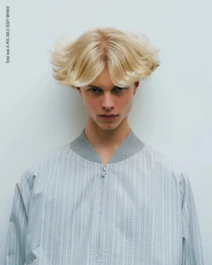 Blond male model with flared parted hair wearing a pale grey textured zip-up jacket against a plain light studio background.
